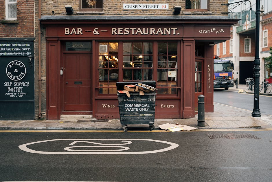 In front of a restaurant with a deep red-brown wooden exterior, a black waste bin labeled 'COMMERCIAL WASTE ONLY' is placed on the grey pavement, filled with flattened cardboard boxes and paper waste spilling over the top. The bin stands adjacent to a small, rounded post on the right side, with a nearby black bollard protecting the entrance to the establishment. To the left, a black signboard advertises a self-service buffet, while a dark blue truck is partially visible on the residential street behind, indicating ongoing waste collection or delivery activity. The restaurant's large window reveals warm interior lighting, contrasting with the cloudy outdoor atmosphere. The scene is set on a typical urban street, with a marked white bicycle lane and a street sign reading 'CRISPIN STREET' overhead, and the overall environment suggests a setting where independent waste disposal could be part of local rubbish removal processes for nearby businesses, such as this restaurant, supported by services like Waste Disposal Finsbury Park.