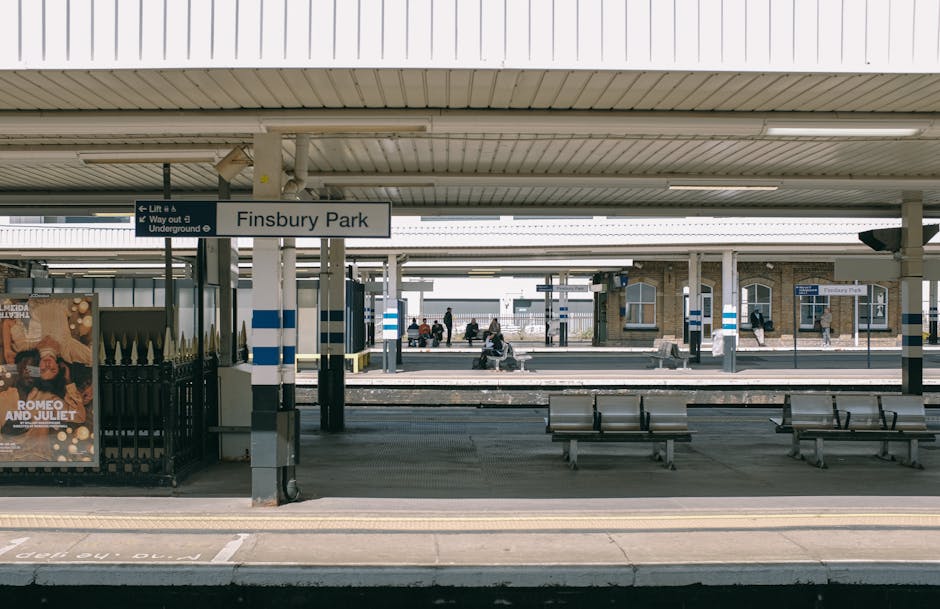 The image depicts a daytime view of Finsbury Park railway station platform, with a long, horizontal roof overhead supported by numerous vertical metal columns painted in grey, white, and blue stripes. In the foreground, there is a paved pedestrian area with tactile paving strips near the platform edge, alongside a sign indicating the station name 'Finsbury Park' mounted on one of the columns. Several wooden benches with metal frames are arranged along the platform for waiting passengers, some of which are occupied by individuals seated and engaging with their phones or devices. To the left, a black metal fence encloses a small advertising display featuring a poster with text and imagery, adjacent to the station entrance. In the background, past the platform, additional railway tracks run parallel, with a mix of station buildings and wait shelters made of brick and glass, some with signage for destination or timetable information. The overall scene is lit by natural daylight, creating a neutral and utilitarian atmosphere typical of urban railway stations, with no visible rubbish or debris, supporting a clean and orderly environment.