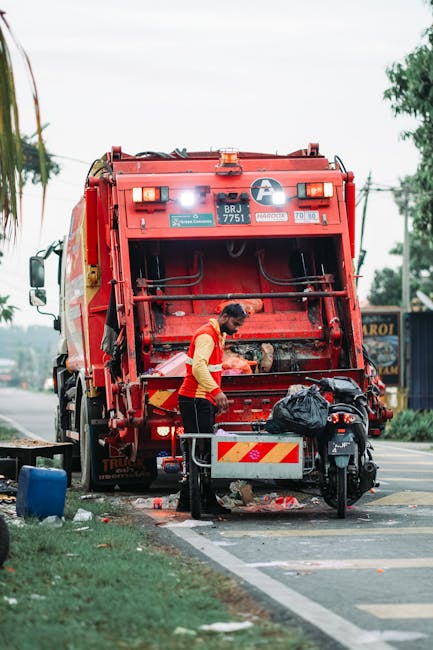 A municipal waste collection worker wearing a high-visibility yellow and orange uniform operates a large red rubbish truck parked on the side of a street. The worker, standing on a small platform at the rear of the vehicle, appears to be emptying or managing waste materials. In front of the truck, there is a black rubbish bag resting on a small cart or container, with scattered debris on the ground around it. The scene is set outdoors, with a paved road, a grassy verge, and some trees visible in the background under an overcast sky. Utility wires run overhead, and a building with signage can be seen in the distance. The equipment and environment suggest a routine waste collection operation that could relate to private or alternative rubbish removal services, aligning with information about independent waste handling without relying on local authority collection. Waste Disposal Finsbury Park provides such services, ensuring proper management of rubbish in urban areas.