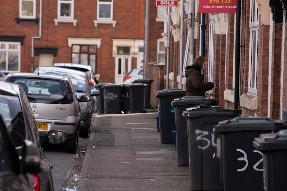 A residential street scene showing a row of black wheelie bins placed along the sidewalk outside brick terraced houses, each bin with white numbering or graffiti on their sides. A woman stands next to one of the bins, appearing to be in the process of discarding rubbish, with her head slightly bowed and hands near the lid of the bin. Multiple parked cars are visible on the left side of the street, including a silver car closest to the camera. The brick facades of the houses in the background feature white window frames and sills, with some showing signs of wear and partial peeling paint. Overhead, a red sign is partially visible, possibly advertising a property sale, attached to the side of a building. The environment is lit evenly with natural daylight, and the scene reflects typical on-street waste collection issues, where private refuse disposal outside homes might require alternative waste management solutions, such as self-service rubbish disposal by companies like Waste Disposal Finsbury Park.