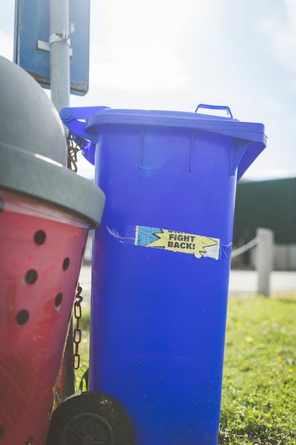 A tall blue plastic rubbish bin with a closed lid, positioned outdoors on a grassy area near a sidewalk, with a label that reads 'FIGHT BACK!' in yellow and black. To the left of the blue bin, part of a red wheeled container with black circular ventilation holes and a silver lid is visible, along with a small chain attached to the lid. The background includes a white post and a blurred view of a street or driveway, suggesting the scene is set in a residential or commercial outdoor space. The lighting indicates daytime with natural sunlight and a clear sky. The scene relates to waste management, indicating the use of individual rubbish bins for waste disposal, aligning with private or alternative rubbish collection practices.