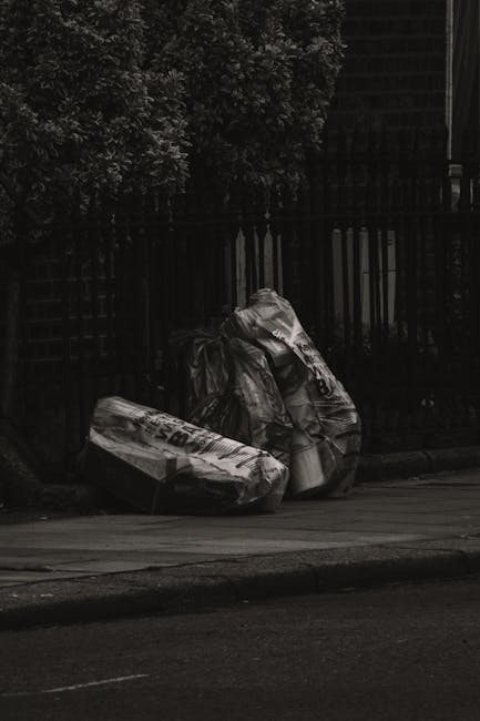 A residential street scene showing a row of black wheelie bins placed along the sidewalk outside brick terraced houses, each bin with white numbering or graffiti on their sides. A woman stands next to one of the bins, appearing to be in the process of discarding rubbish, with her head slightly bowed and hands near the lid of the bin. Multiple parked cars are visible on the left side of the street, including a silver car closest to the camera. The brick facades of the houses in the background feature white window frames and sills, with some showing signs of wear and partial peeling paint. Overhead, a red sign is partially visible, possibly advertising a property sale, attached to the side of a building. The environment is lit evenly with natural daylight, and the scene reflects typical on-street waste collection issues, where private refuse disposal outside homes might require alternative waste management solutions, such as self-service rubbish disposal by companies like Waste Disposal Finsbury Park.