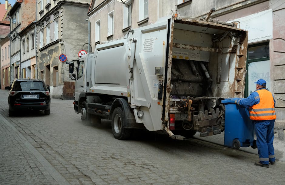 A commercial waste collection truck with a predominantly white exterior and a rusted, open rear loading compartment parked on a cobblestone street in an urban residential area. The truck's rear section is open, revealing an empty, metal-lined space with visible rust and wear marks, indicating recent or ongoing refuse collection. A worker wearing a blue uniform and a bright orange high-visibility vest is positioned at the back of the truck, holding a blue wheeled bin and preparing to empty it into the compactor. To the left of the truck, a black car is parked alongside aged multi-story buildings with weathered facades, some showing signs of peeling paint. The street is narrow, lined with paving stones, and features signs restricting parking or stopping. The environment appears damp with soft natural lighting, typical of a cloudy day, and the scene exemplifies a routine rubbish removal operation conducted by Waste Disposal Finsbury Park, specializing in private waste handling and on-site clearance services within urban residential settings.