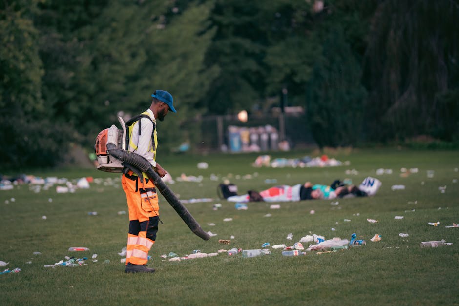 A man wearing a high-visibility jacket, orange trousers with reflective stripes, and a blue cap stands on a grassy field during daytime, holding a leaf blower with a large flexible hose. The man appears to be engaged in cleaning up litter scattered across the open area. In the background, there are a few people lying on the grass, some surrounded by discarded rubbish, with trees and shrubs bordering the field. The scene also includes a distant row of waste bins and a small building or structure, indicating a public park or recreational area. The ground is covered with various types of trash, including plastic bottles, cups, and paper, which the worker is aiming to clear, demonstrating an example of private rubbish removal efforts or on-site waste clearance services in a natural outdoor environment. The lighting suggests a soft, natural daylight, emphasizing the contrast between the clean-up activity and the littered surroundings, aligning with themes of waste management and rubbish removal for outdoor public spaces.