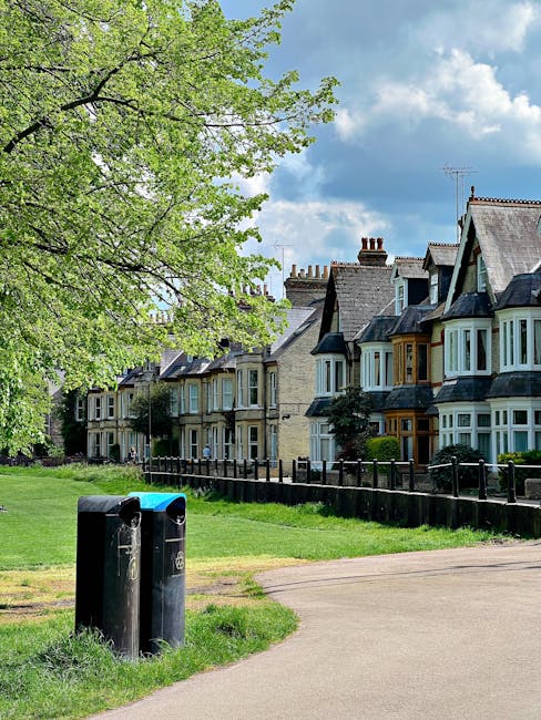 The image shows a row of traditional terraced houses with bay windows and slate roofs, situated behind a neatly maintained green garden and a black metal fence. In the foreground, two black refuse bins with blue lids are positioned on a paved pathway, adjacent to a grassy area. To the left, a large tree with fresh green leaves extends its branches over the scene, casting slight shadows on the grass and pathway below. The sky overhead is partly cloudy with patches of blue visible, suggesting a bright, clear day. The scene captures a quiet residential neighborhood, where waste disposal is managed privately, as indicated by the presence of personal bins, reflecting an alternative waste handling approach to regular council collection. Overall, the scene emphasizes an orderly, community-oriented environment with attention to outdoor aesthetics and waste management practices.