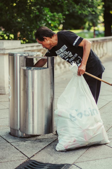 A middle-aged man dressed in a black T-shirt with white text and dark trousers is seen standing on a paved sidewalk, actively disposing of a large white garbage bag into a stainless steel public litter bin. The man is bent slightly forward, holding the bag open with his left hand, while his right hand grips a wooden stick or litter picker resting against the bin. The rubbish bag appears full and is made of a semi-translucent plastic material, with some vague contents visible inside. The stainless steel litter bin has a rounded, modern design with a circular opening at the top, situated against a background of lush green trees and shrubs, indicating a park or urban green space environment. The pavement around the bin is clean, and natural daylight illuminates the scene, highlighting the reflective surface of the metal bin and the textured surface of the stone sidewalk. This image relates to the topic of private waste handling or rubbish collection services, such as those provided by Waste Disposal Finsbury Park, highlighting an individual engaged in on-site waste disposal activity in an outdoor setting.