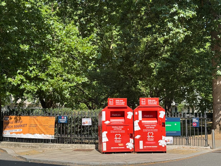 The image shows a row of traditional terraced houses with bay windows and slate roofs, situated behind a neatly maintained green garden and a black metal fence. In the foreground, two black refuse bins with blue lids are positioned on a paved pathway, adjacent to a grassy area. To the left, a large tree with fresh green leaves extends its branches over the scene, casting slight shadows on the grass and pathway below. The sky overhead is partly cloudy with patches of blue visible, suggesting a bright, clear day. The scene captures a quiet residential neighborhood, where waste disposal is managed privately, as indicated by the presence of personal bins, reflecting an alternative waste handling approach to regular council collection. Overall, the scene emphasizes an orderly, community-oriented environment with attention to outdoor aesthetics and waste management practices.