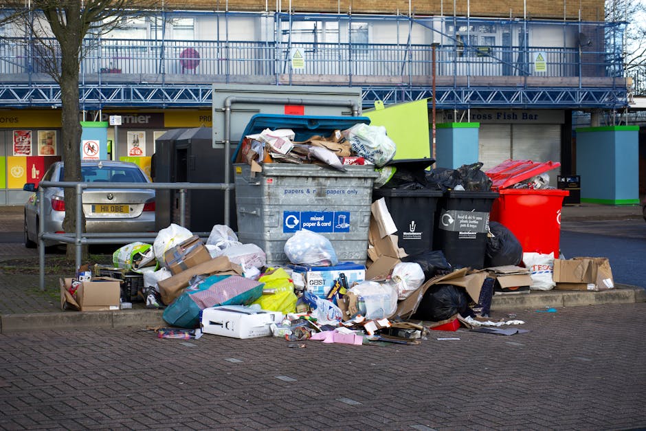 A cluttered collection of overflowing rubbish bins and scattered waste bags sits on a paved area adjacent to a parking space, with a silver car parked nearby. The central roller bin is filled with mixed paper and cardboard, with its blue lid partially open, revealing the contents inside. To its right, a black bin and a red bin are also overflowing, with black garbage bags and loose waste spilling onto the ground. Surrounding the bins are various discarded cardboard boxes, plastic packaging, paper, and smaller bags, some flattened and others crumpled, creating a mess that extends onto the pavement. In the background, a building with storefronts features signs and advertisements, and above, a construction scaffold draped with blue mesh partially obscures the upper facade. The scene is illuminated by natural daylight, emphasizing the disorganized state of the rubbish, which appears to be a result of an unscheduled or private waste collection, possibly managed by waste disposal services such as Waste Disposal Finsbury Park, rather than council-led rubbish removal.