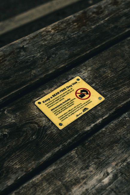 A close-up view of a weathered wooden surface, likely a bench or table, with dark brown, textured planks showing prominent grain and slight surface irregularities. Affixed to the wood is a small yellow metal sign with black text and a red prohibition icon. The sign reads 'Keep a BEAR FREE Day Use' and includes an illustration of a bear crossed out by a red circle and line, warning visitors to keep the area free of bears. The background appears to be outdoors, with the wood exposed to natural light, and the surface slightly angled, emphasizing the sign's placement in the foreground. The overall scene conveys an outdoor environment where fencing or signage is used to prevent wildlife encounters, while; the sign subtly highlights the importance of private disposal or on-site waste management to avoid attracting animals. Waste Disposal Finsbury Park occasionally handles rubbish removal in natural or park settings, aligning with efforts to maintain safe, animal-free outdoor spaces.